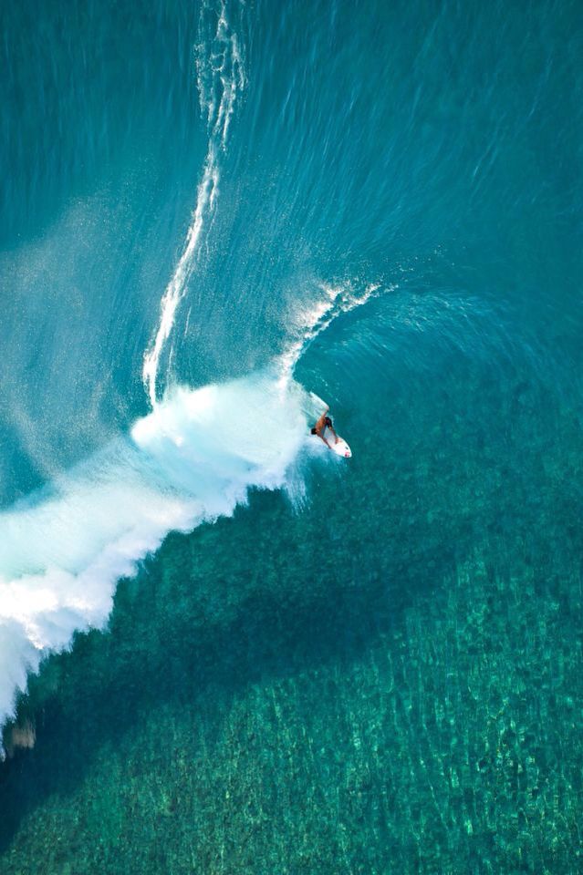 Aerial view of a surfer riding a wave over clear turquoise water, leaving a white trail behind. The ocean floor is visible through the transparent, shallow water.