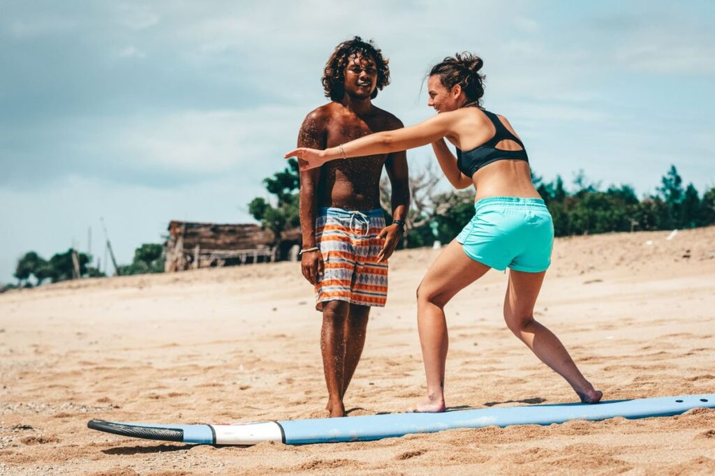 A woman practices surfing on the sand while a man stands nearby, guiding her. They are on a sandy beach with trees and a small wooden structure in the background, both dressed in swimwear.