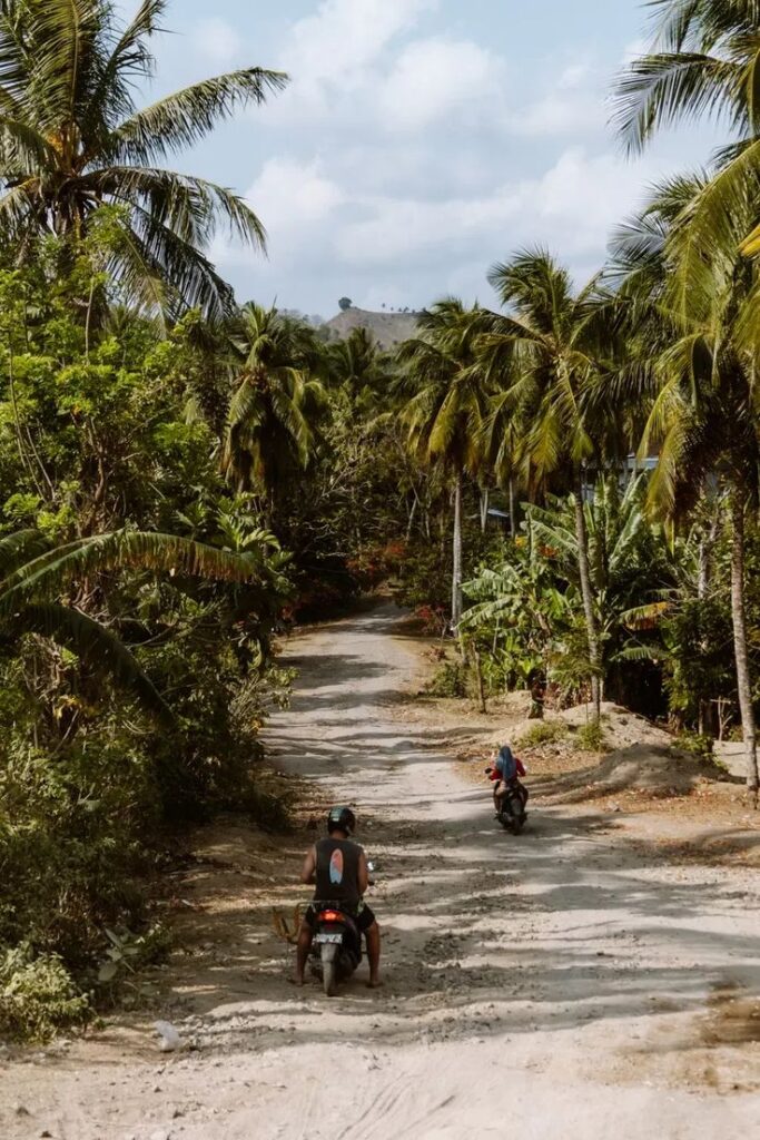 Two people ride motorbikes down a dirt road surrounded by lush green palm trees and tropical vegetation under a partly cloudy sky.