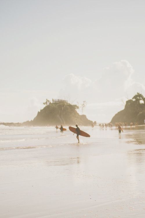 A surfer carrying a longboard walks along a sandy beach with gentle waves and rocky outcrops in the distance, under a bright, hazy sky. Other people are seen enjoying the beach in the background.