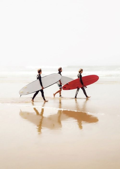 Three surfers in wetsuits walk along a sandy beach, each carrying a surfboard—two white, one red—with their reflections visible on the wet sand and gentle ocean waves in the background.