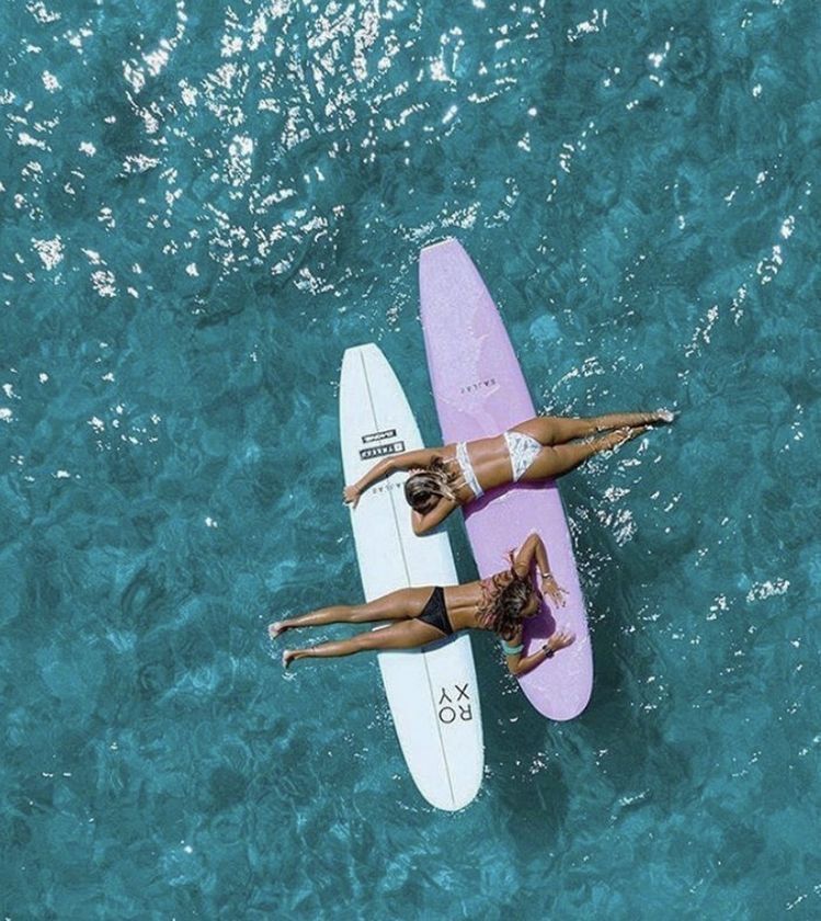 Two women in swimsuits lie on surfboards, one white and one pink, floating on clear blue water. They are facing down and reaching toward each other, surrounded by rippling waves.