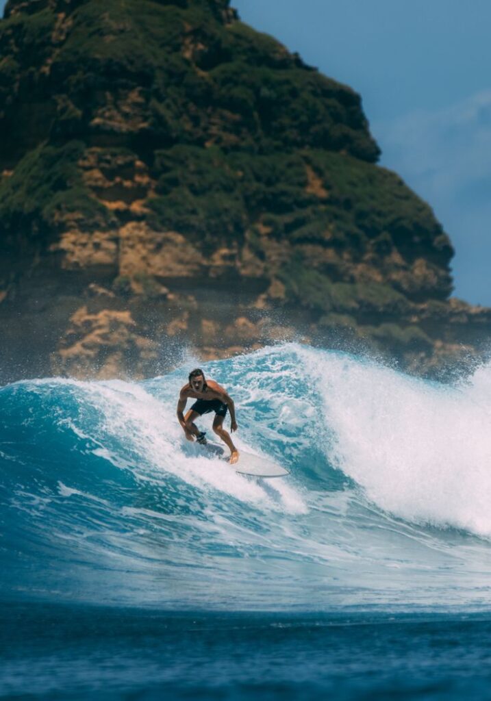 A surfer rides a blue wave in the ocean with a rocky, green-covered cliff in the background under a clear sky.