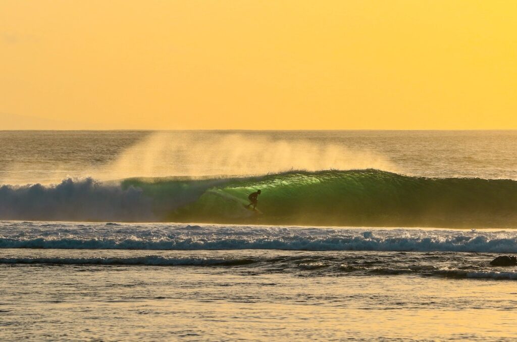 A surfer rides inside a green ocean wave at sunset, with golden sunlight illuminating the sky and water, creating a serene and vibrant scene.