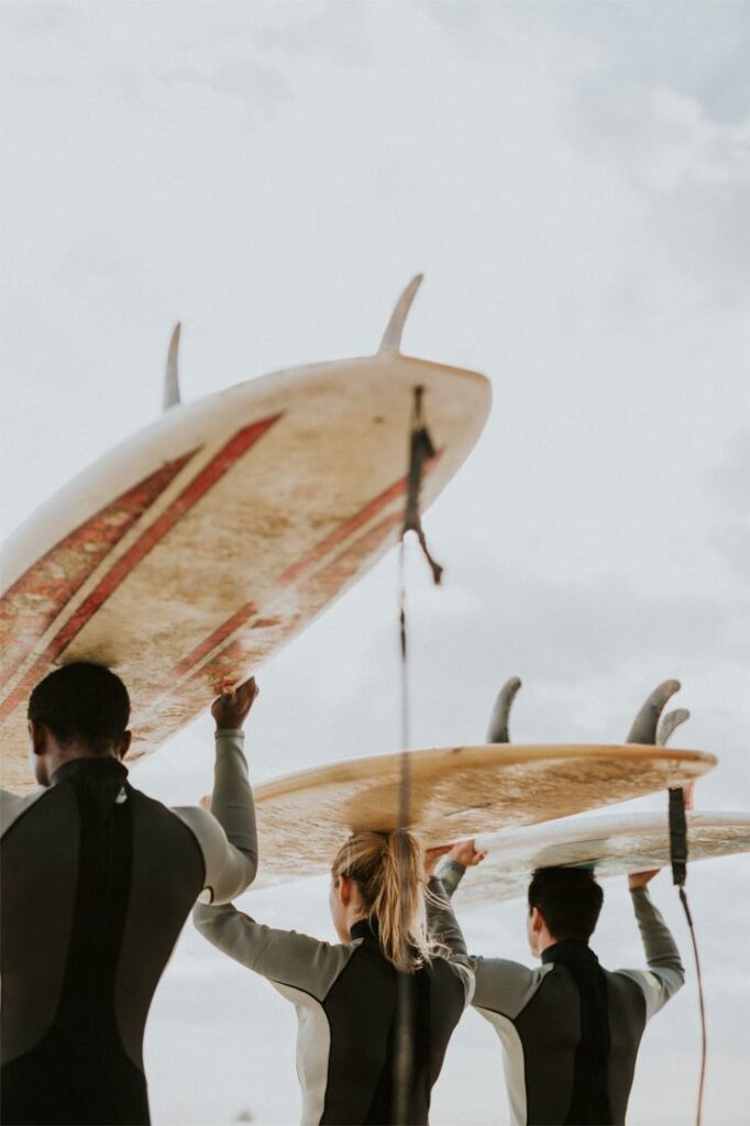 Three people in wetsuits carry surfboards above their heads, walking towards the ocean under a cloudy sky. The photo is taken from behind, capturing the surfboards and backs of the surfers.