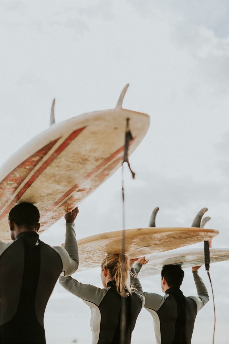 Three people in wetsuits carry surfboards above their heads, walking towards the ocean under a cloudy sky. The photo is taken from behind, capturing the surfboards and backs of the surfers.
